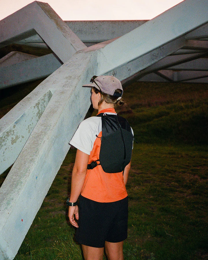 Person wearing an orange shirt and black backpack standing under a large metal structure.