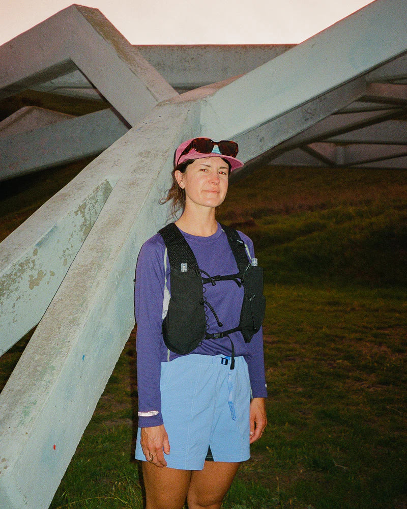 Person wearing a purple long-sleeve shirt and blue shorts with a black backpack, standing under a large metal structure.