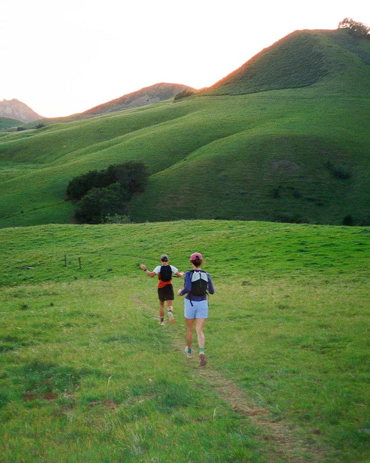 Two people running on a grassy hill with rolling green hills in the background.