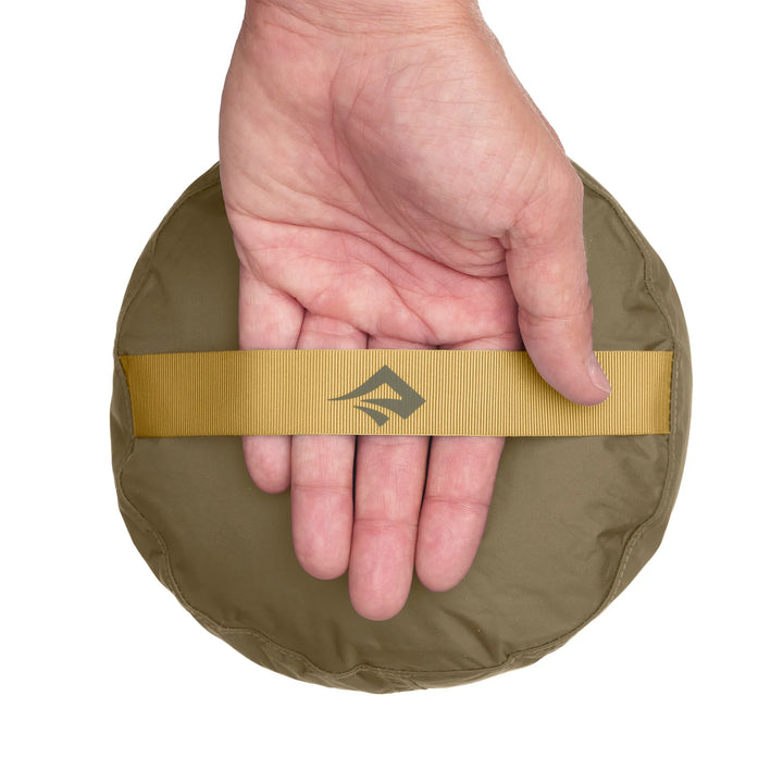 Hand holding a khaki-colored round object with a gold band featuring a logo on a white background