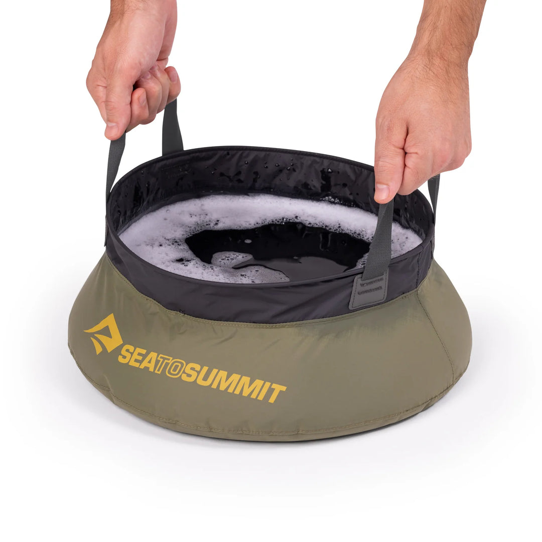 Portable sink with soapy water inside, held by a person, on a white background. Sea to Summit logo visible.