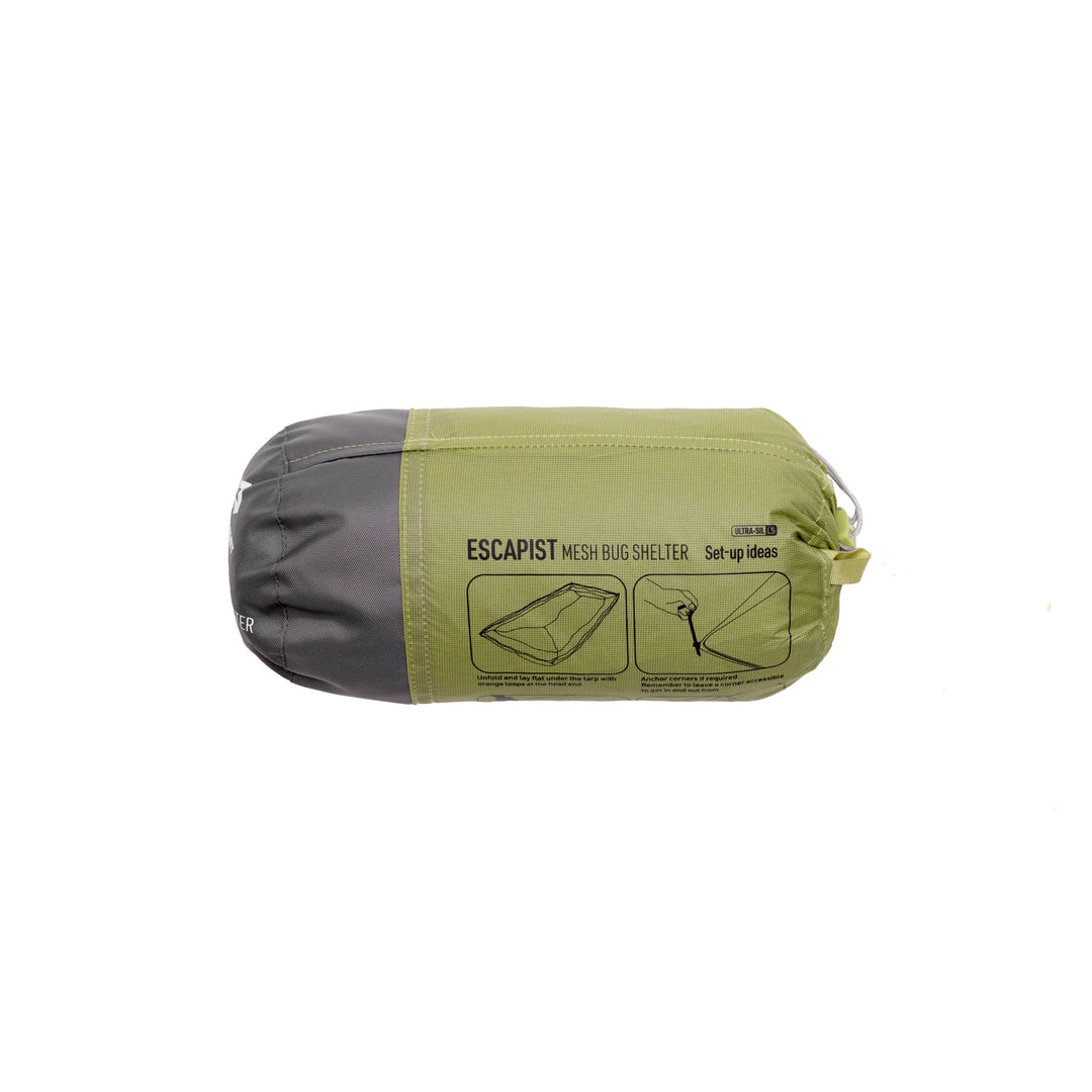 A packed mesh bug shelter bag, shown horizontally against a plain white background. The bag is olive green on the outside with a dark gray interior visible at the left end, and features printed black text and diagrams on the front.