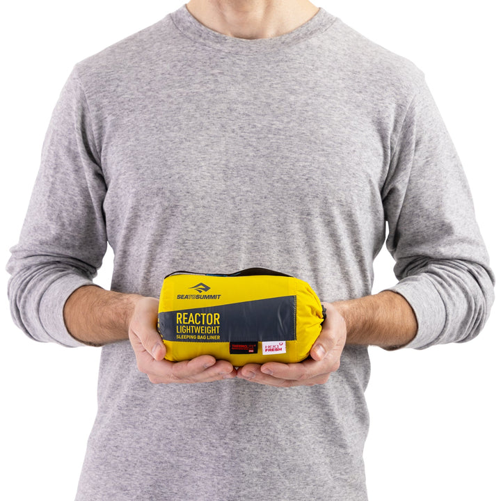 Person holding a yellow sleeping bag liner in stuff sack on a white background