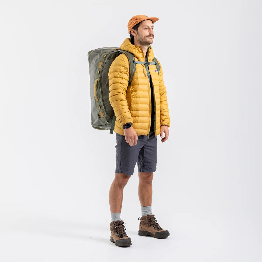 A man wearing a yellow puffer jacket, dark gray shorts, brown hiking boots, and an orange cap, carrying a large gray hiking backpack against a plain white studio background.