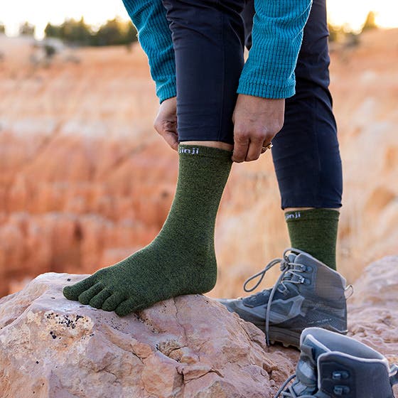 Person wearing green toe socks with blue sweater and dark pants, standing on a rock with a natural landscape background