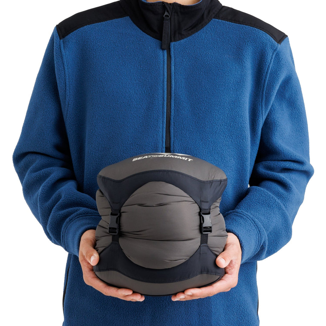 Person wearing a blue fleece jacket and holding a sleeping bag in a grey stuff sack