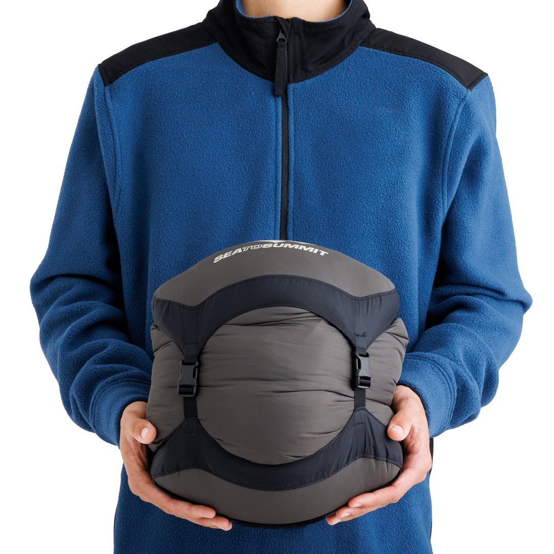 Person wearing a blue fleece jacket and holding a sleeping bag in a grey stuff sack