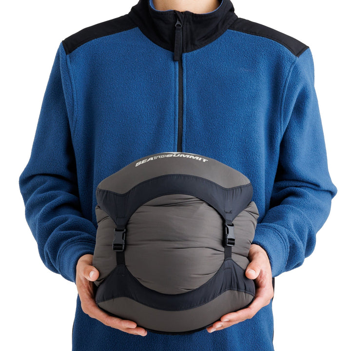 Person wearing a blue fleece jacket and holding a sleeping bag in a grey stuff sack