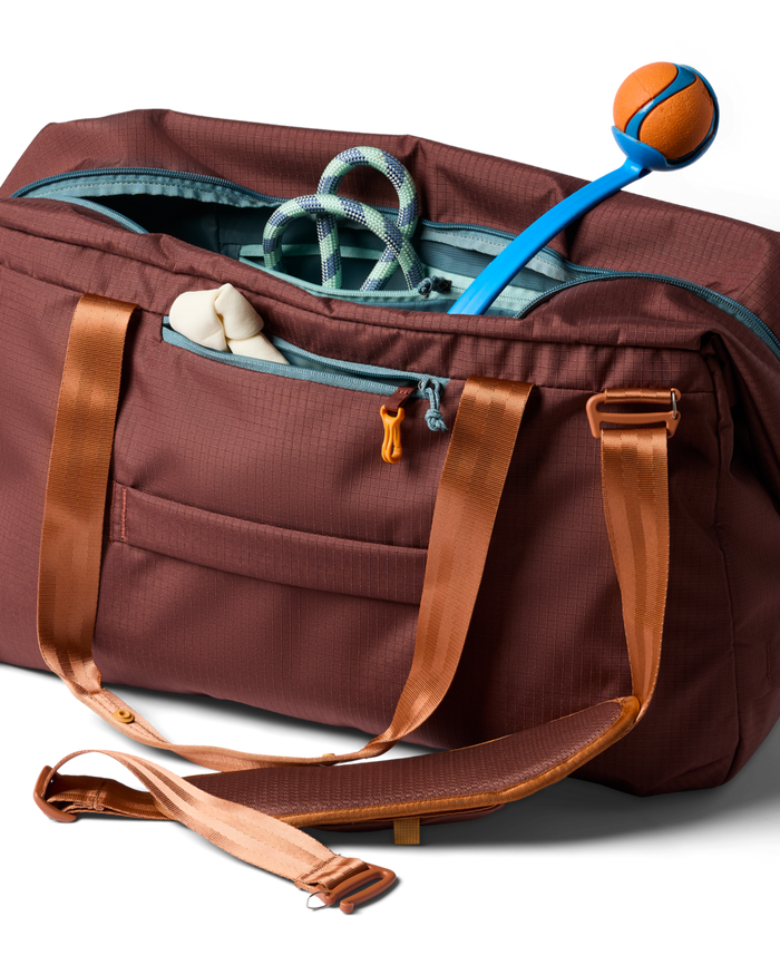 Brown duffel bag with brown straps containing a rope, towel, and blue and orange ball on a white background.