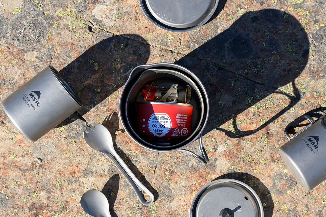 Camping cookware set with pots, pans, and a lighter on a textured surface