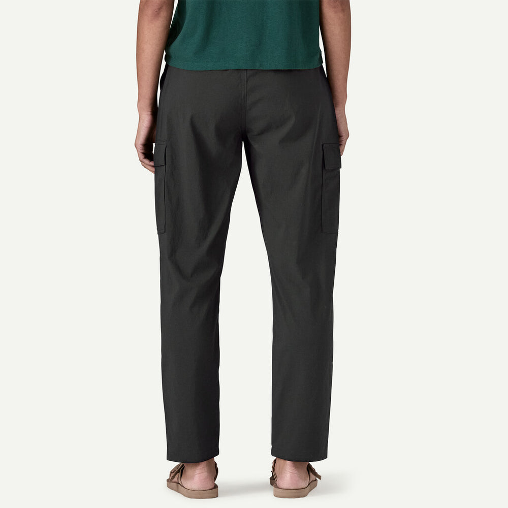 Rear view of a person wearing dark charcoal cargo pants with side flap pockets, paired with a green top and tan sandals, against a light gray studio background.