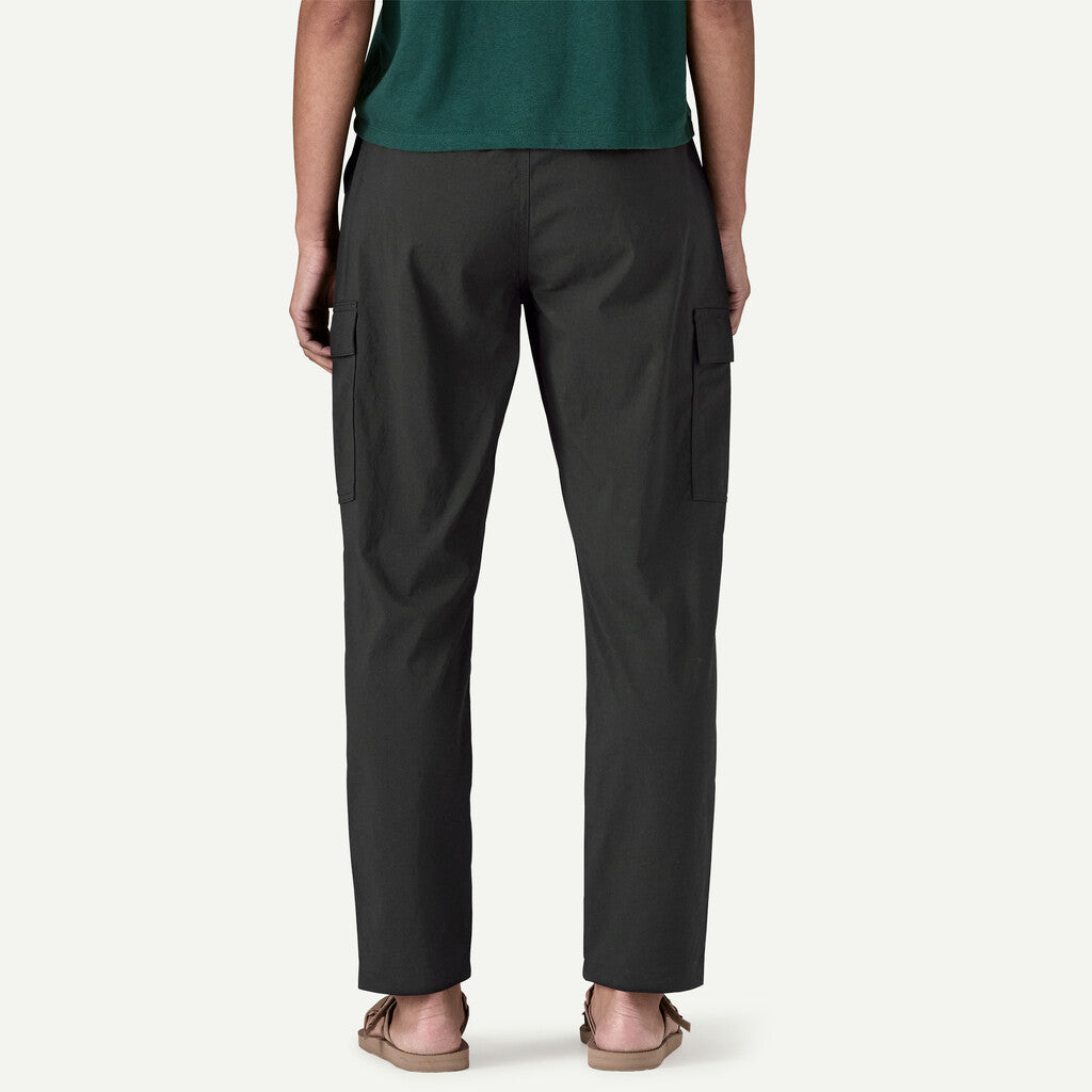 Rear view of a person wearing dark charcoal cargo pants with side flap pockets, paired with a green top and tan sandals, against a light gray studio background.