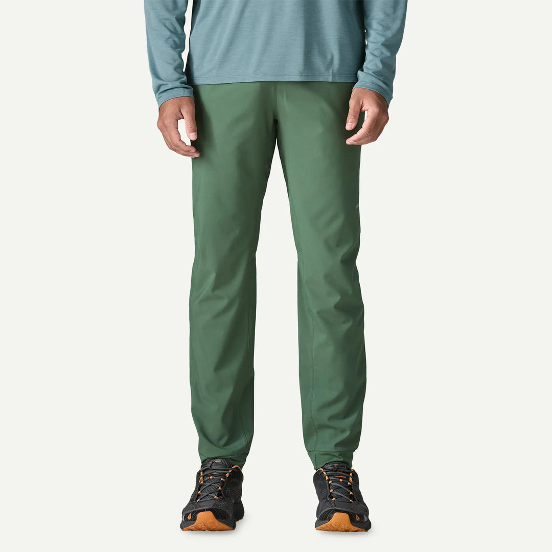 A person wearing slim-fit green pants, a muted blue-green long-sleeve top, and black hiking shoes with orange soles, shown from the waist down against a plain off-white studio background.