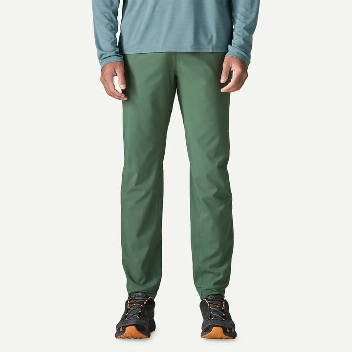 A person wearing slim-fit green pants, a muted blue-green long-sleeve top, and black hiking shoes with orange soles, shown from the waist down against a plain off-white studio background.