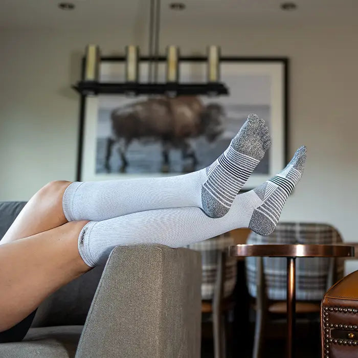 Person wearing light gray socks with dark gray patterns, sitting on a couch in a room with a chandelier and framed picture.