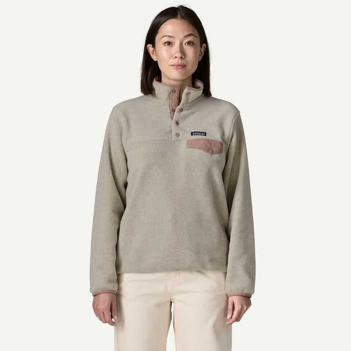 A woman modeling a Patagonia quarter-zip fleece pullover in a light taupe color with a dusty rose pocket and trim, paired with cream pants, against a solid off-white background.