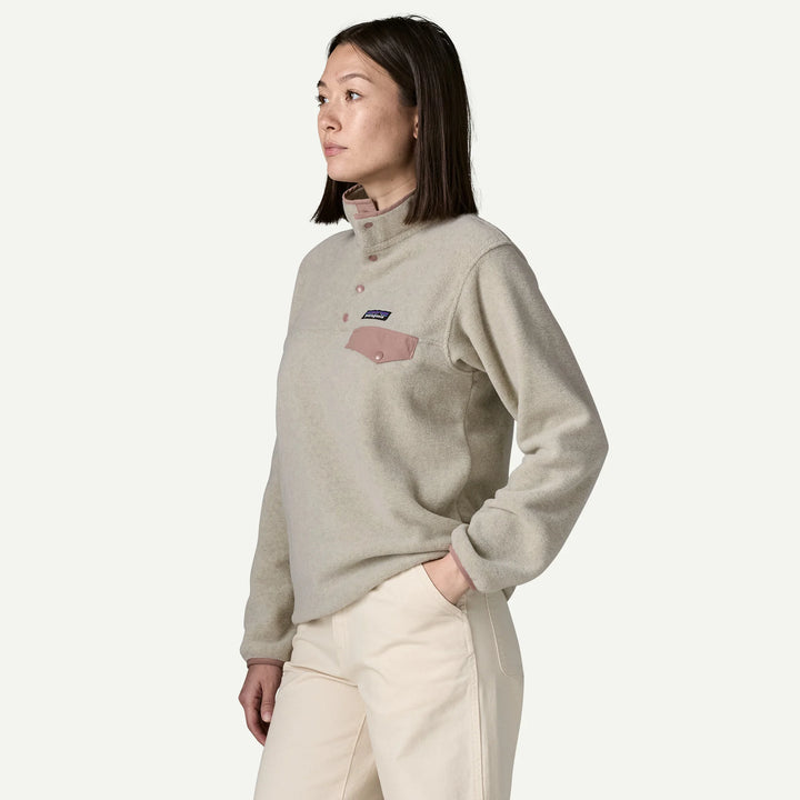 A woman wearing a light beige Patagonia fleece pullover with a pink chest pocket and buttons, paired with cream pants, photographed against a clean off-white studio background.
