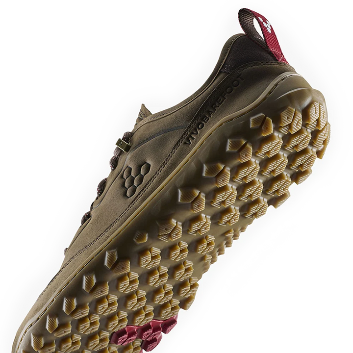 A close-up of a rugged outdoor shoe sole showing deep lugs and a tan rubber tread, with a brown upper and a small red pull tab visible at the heel.