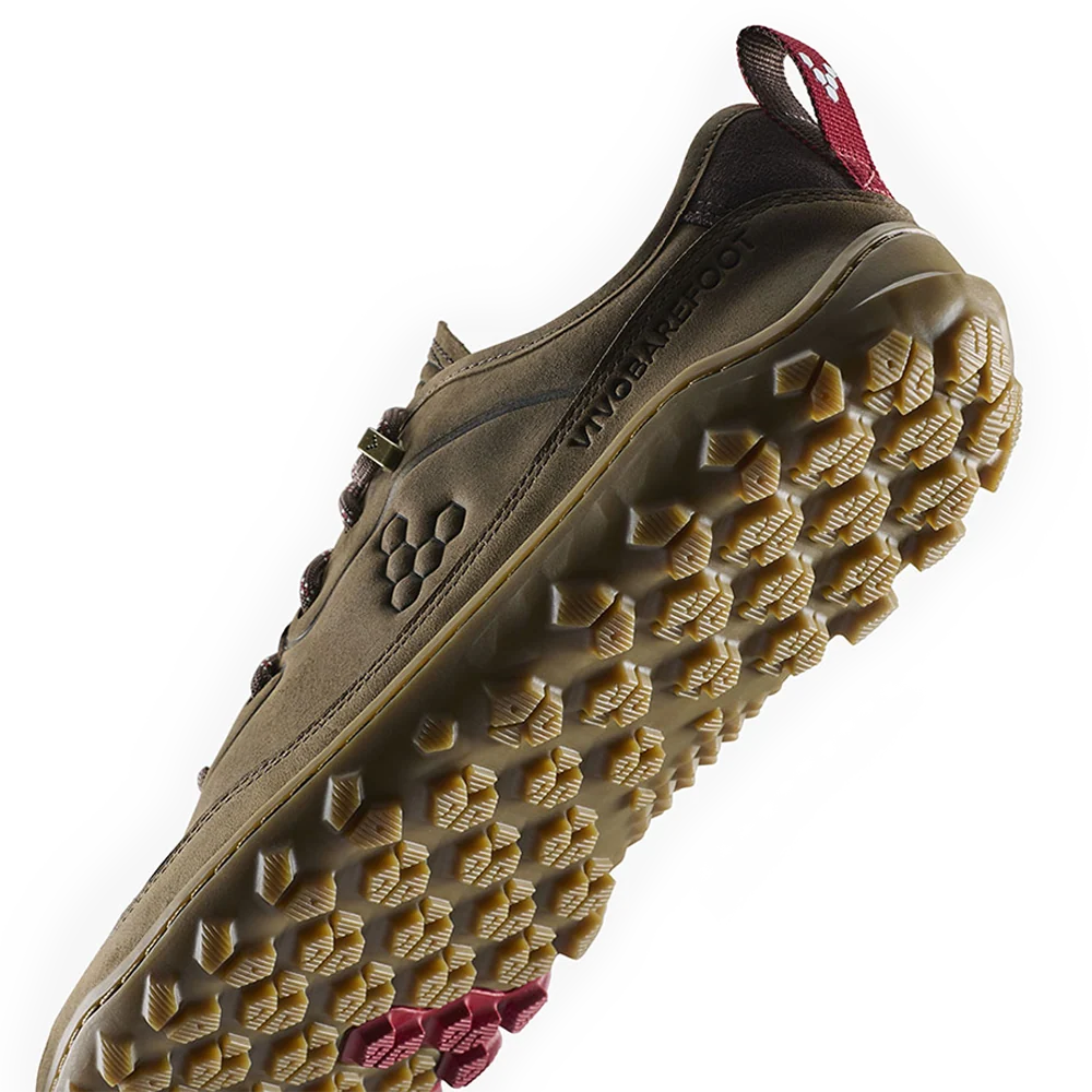 A close-up of a rugged outdoor shoe sole showing deep lugs and a tan rubber tread, with a brown upper and a small red pull tab visible at the heel.