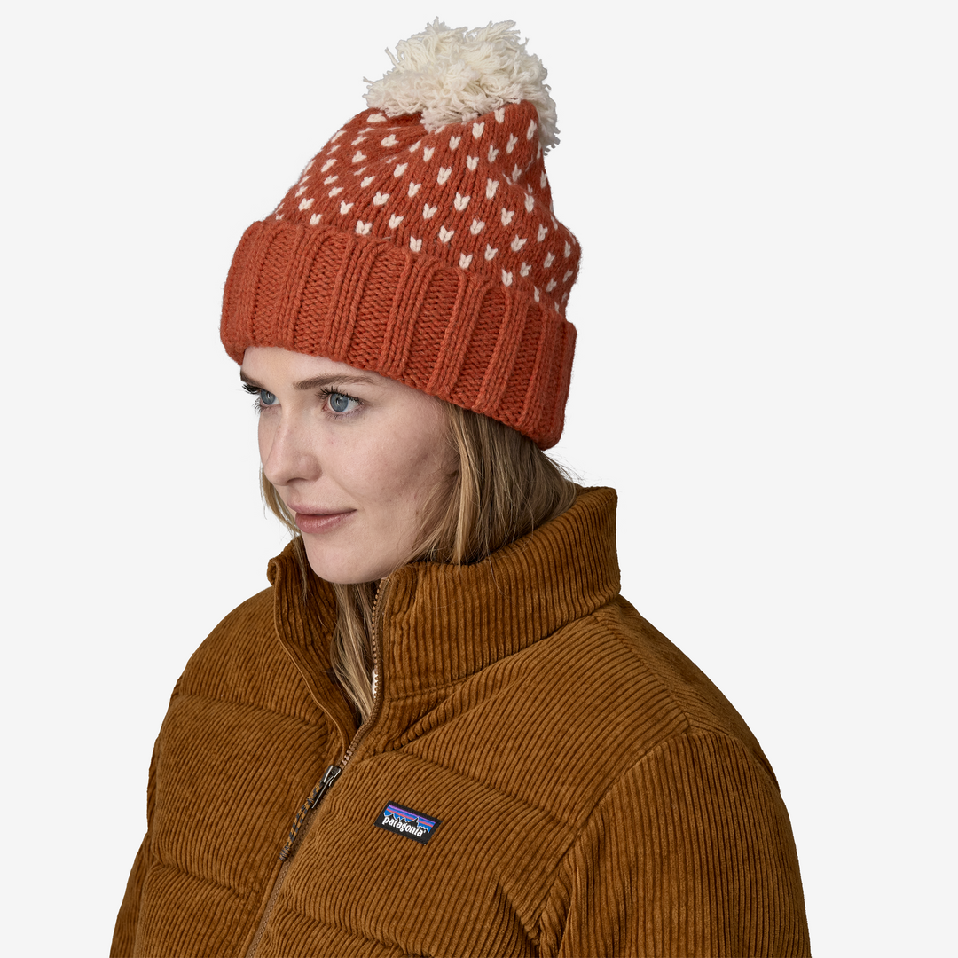 Woman wearing a orange knit beanie with white pattern and brown jacket on a white background