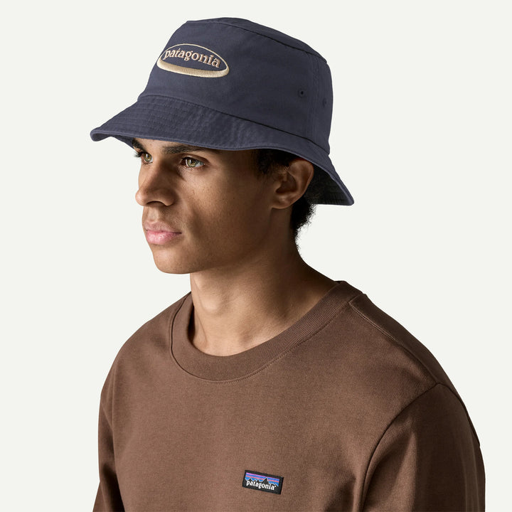 A man wearing a navy blue Patagonia bucket hat and a brown Patagonia crewneck sweatshirt, shown in profile against a light off-white background.