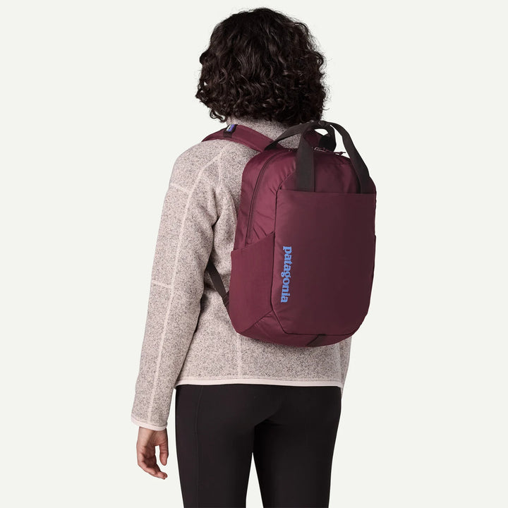 A woman shown from the back wearing a Patagonia backpack in burgundy. The backpack has a vertical Patagonia logo in blue on the side and dark gray handles.