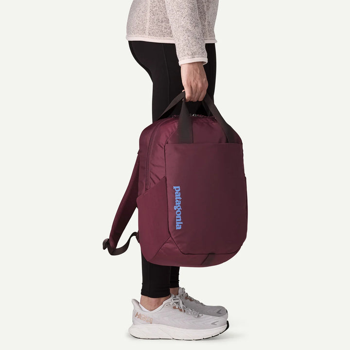 A person wearing a light gray long-sleeve top, black leggings, and gray sneakers holds a Patagonia backpack in a side profile view. The backpack is a deep plum purple with black handles and a light blue Patagonia logo on the side.