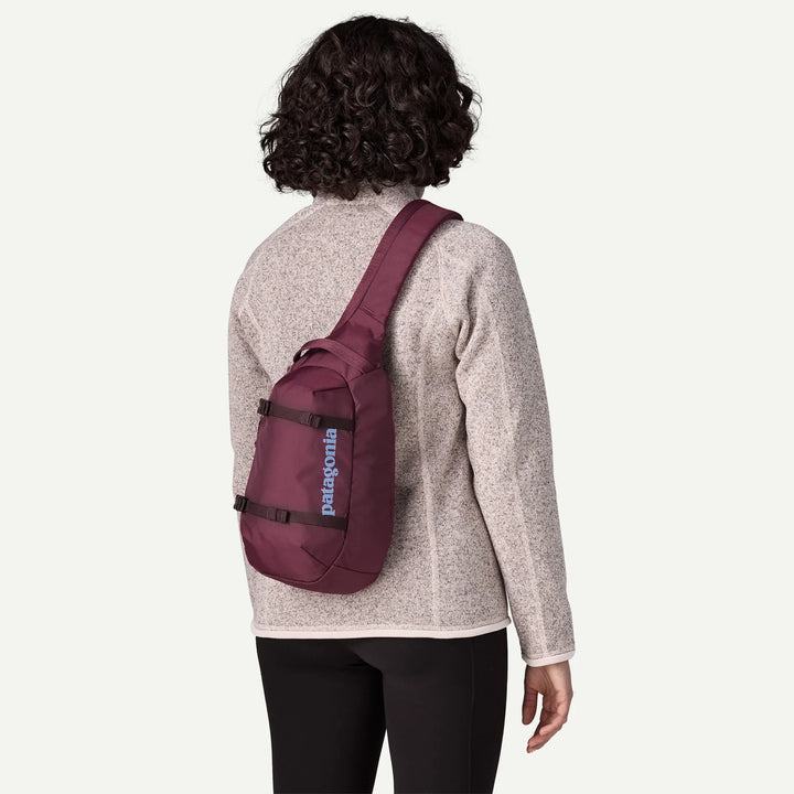 A person shown from the back wearing a Patagonia sling backpack in burgundy, paired with a light heather gray fleece and black pants, against a solid off-white background.