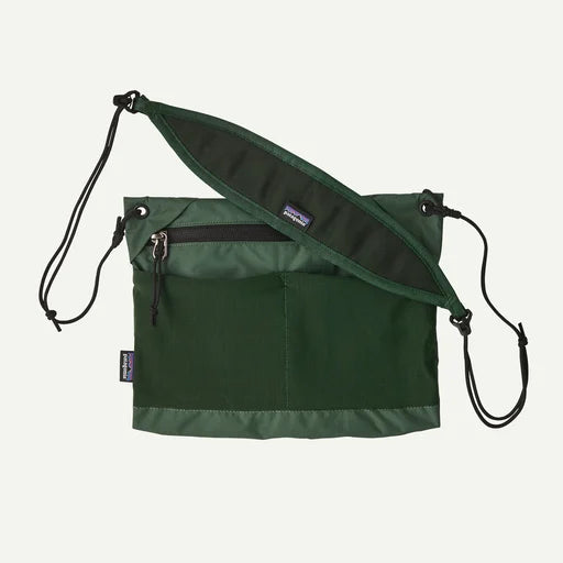 A dark green Patagonia shoulder bag with a wide, padded black shoulder strap laid diagonally across the front. The bag features a front zippered pocket and a vertical front pocket, shown against a plain white background.