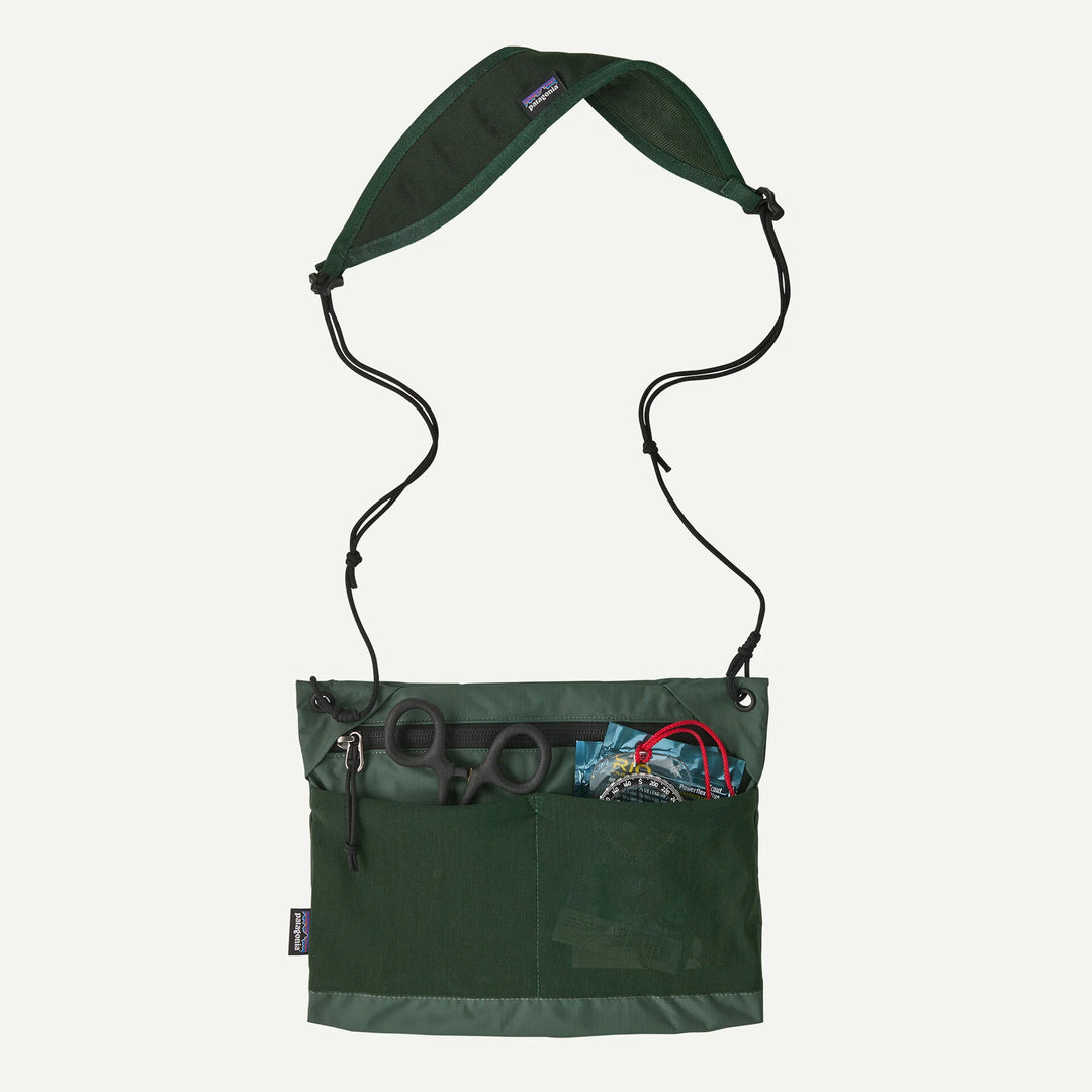 A dark green Patagonia shoulder bag with a black adjustable strap, shown against a light off-white background. The front pocket is open, displaying scissors and other small accessories.