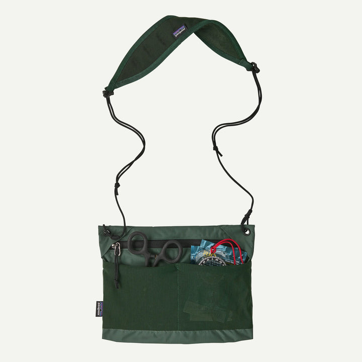 A dark green Patagonia shoulder bag with a black adjustable strap, shown against a light off-white background. The front pocket is open, displaying scissors and other small accessories.