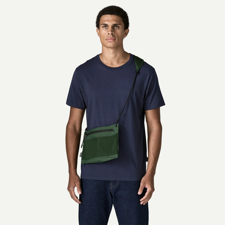 A man wearing a navy blue t-shirt and dark jeans models a green crossbody sling bag with a black strap against a light off-white background.