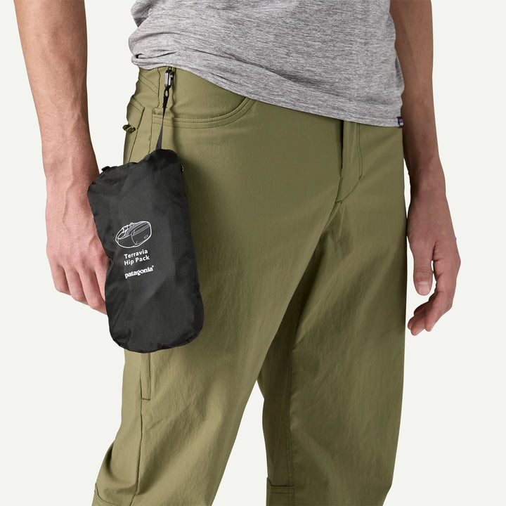 Person wearing green pants holding a black pack with visible branding on a white background