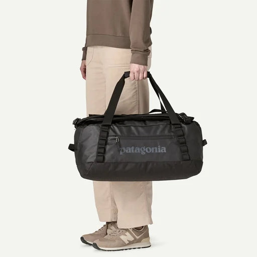 A person wearing beige pants and brown shoes holds a black Patagonia duffel bag with a gray logo on the front, against a plain light background.