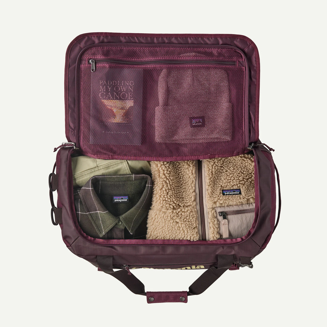An open Patagonia backpack shown from above, filled with travel items including a green Patagonia shirt, a beige fleece jacket, and a zippered pouch. The backpack interior is burgundy, and the items are arranged neatly against a light off-white background.