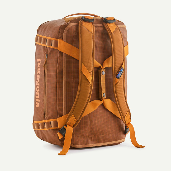 A Patagonia backpack in brown and orange, shown from the back on a clean off-white background. The backpack features a brown textured body with orange webbing straps, a vertical Patagonia logo on the side, and a small blue brand tag on the upper right.