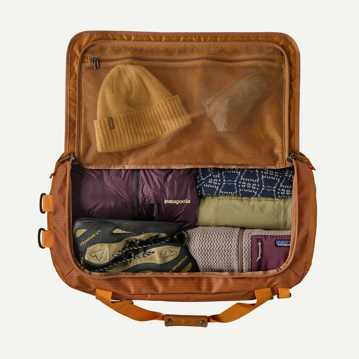 An open Patagonia backpacking suitcase shown from above, packed with travel gear including a tan beanie, purple jacket, patterned shirt, hiking boots, and a knit hat, on a light gray background.
