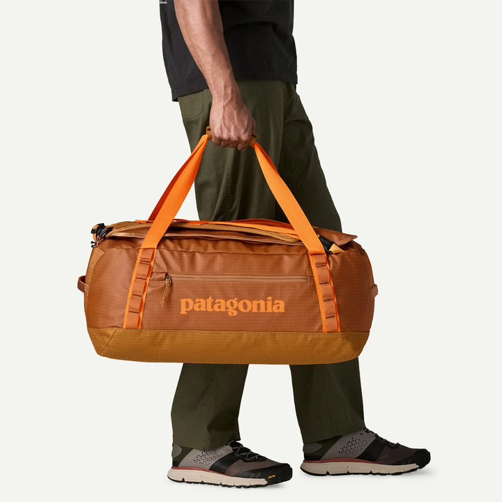 A person wearing olive green pants and black sneakers carries a large Patagonia duffel bag in orange and brown. The person is cropped from the chest down, standing against a clean off-white studio background.