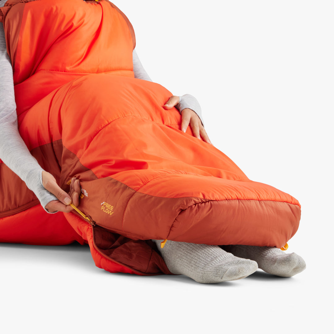 Person sitting up in a orange sleeping bag with their arms and feet sticking out through open zips