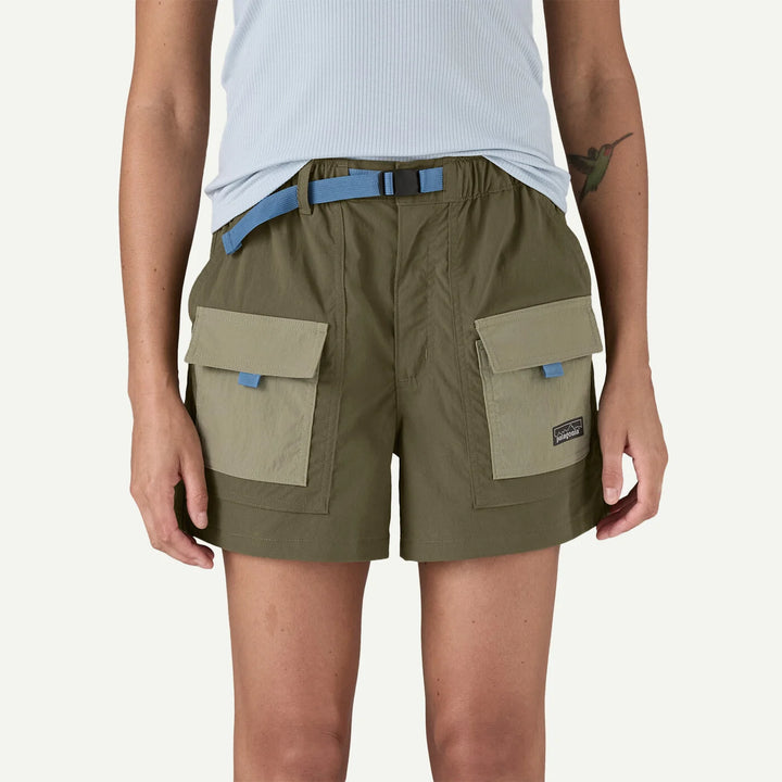A model wearing olive green cargo shorts with light beige flap pockets and a light blue belt, paired with a white ribbed top against a light off-white background.