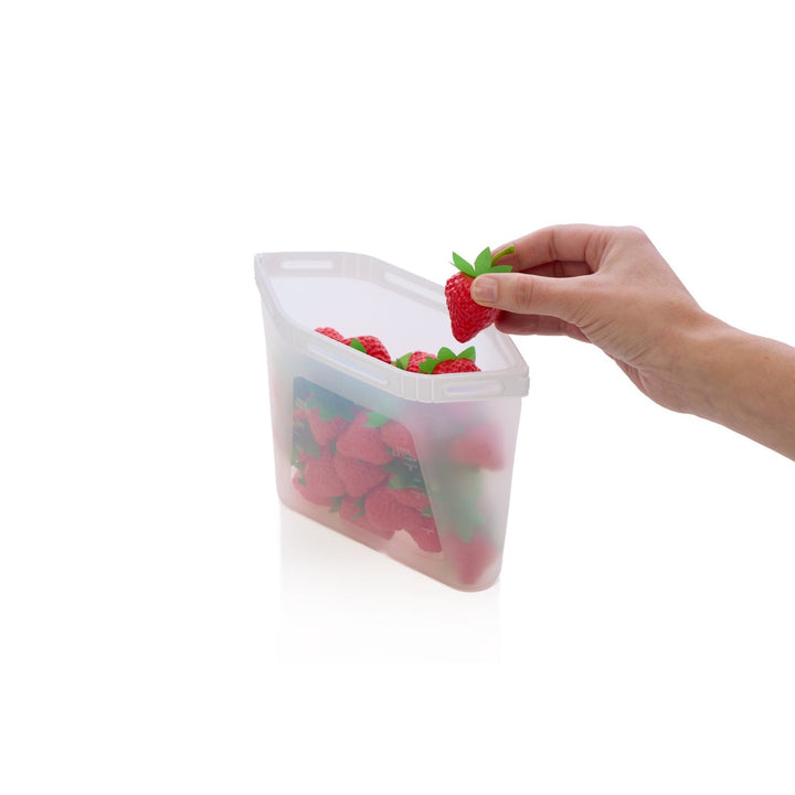 Hand placing a strawberry into a silicone food storage bag on a white background