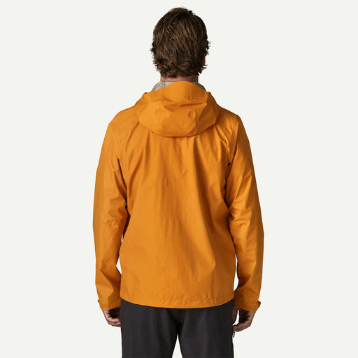 A man viewed from behind wearing a bright orange hooded jacket and dark pants, standing against a plain light gray studio background.