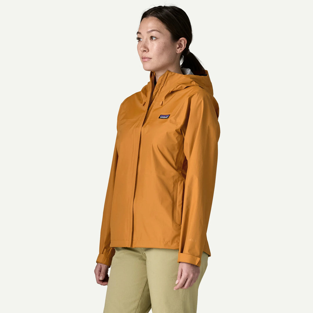A woman wearing a Patagonia hooded jacket in a warm ochre orange color, paired with khaki pants, shown in profile against a light off-white background.