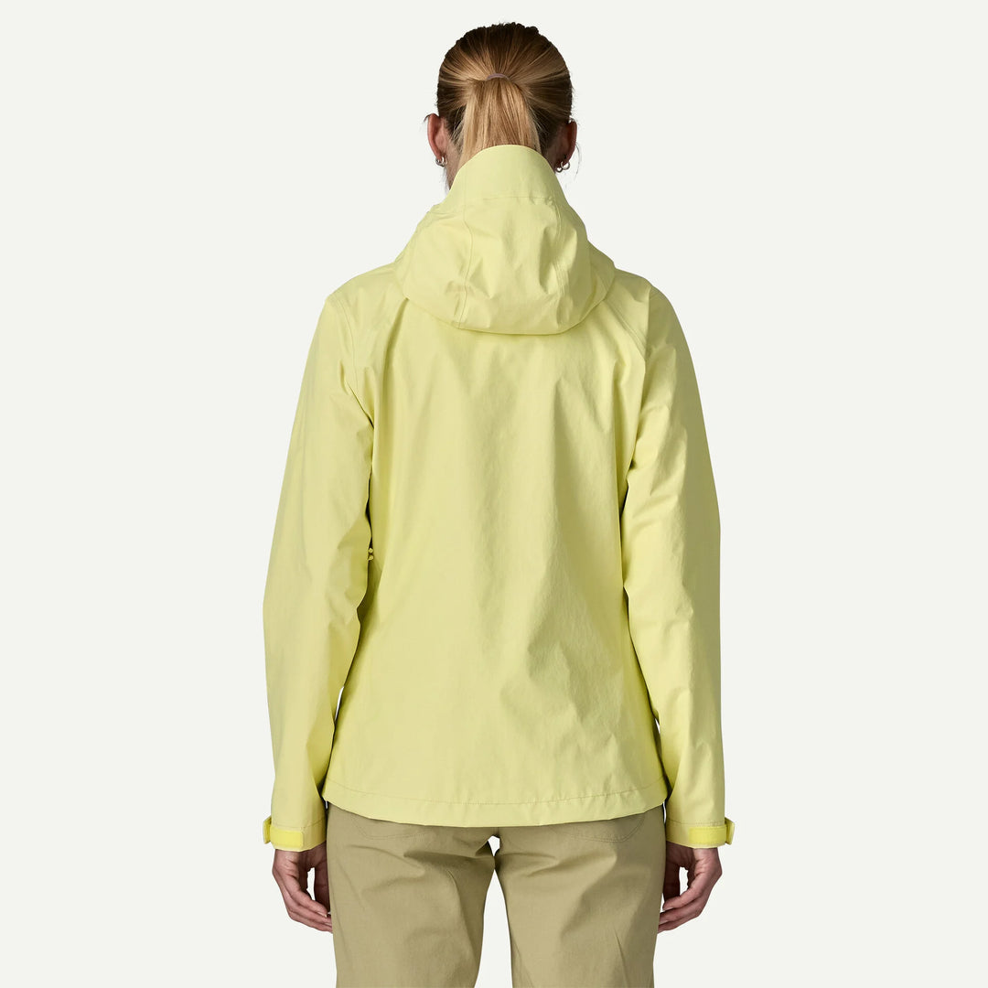 A woman viewed from behind wearing a light yellow hooded jacket and tan pants against a plain light gray studio background.