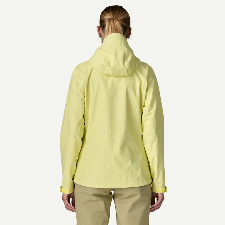 A woman viewed from behind wearing a light yellow hooded jacket and tan pants against a plain light gray studio background.
