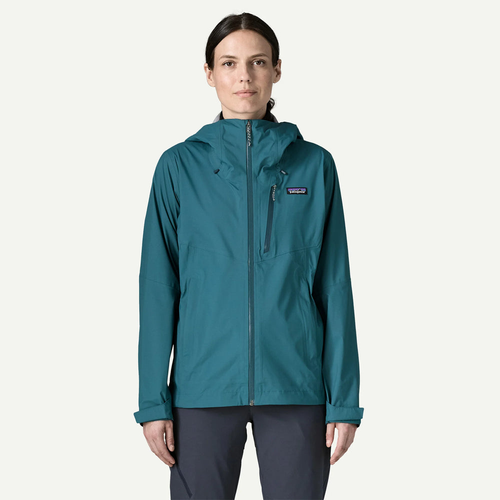 A woman modeling a teal Patagonia hooded jacket with a full front zipper and a zippered chest pocket, paired with dark gray pants, against a light off-white background.