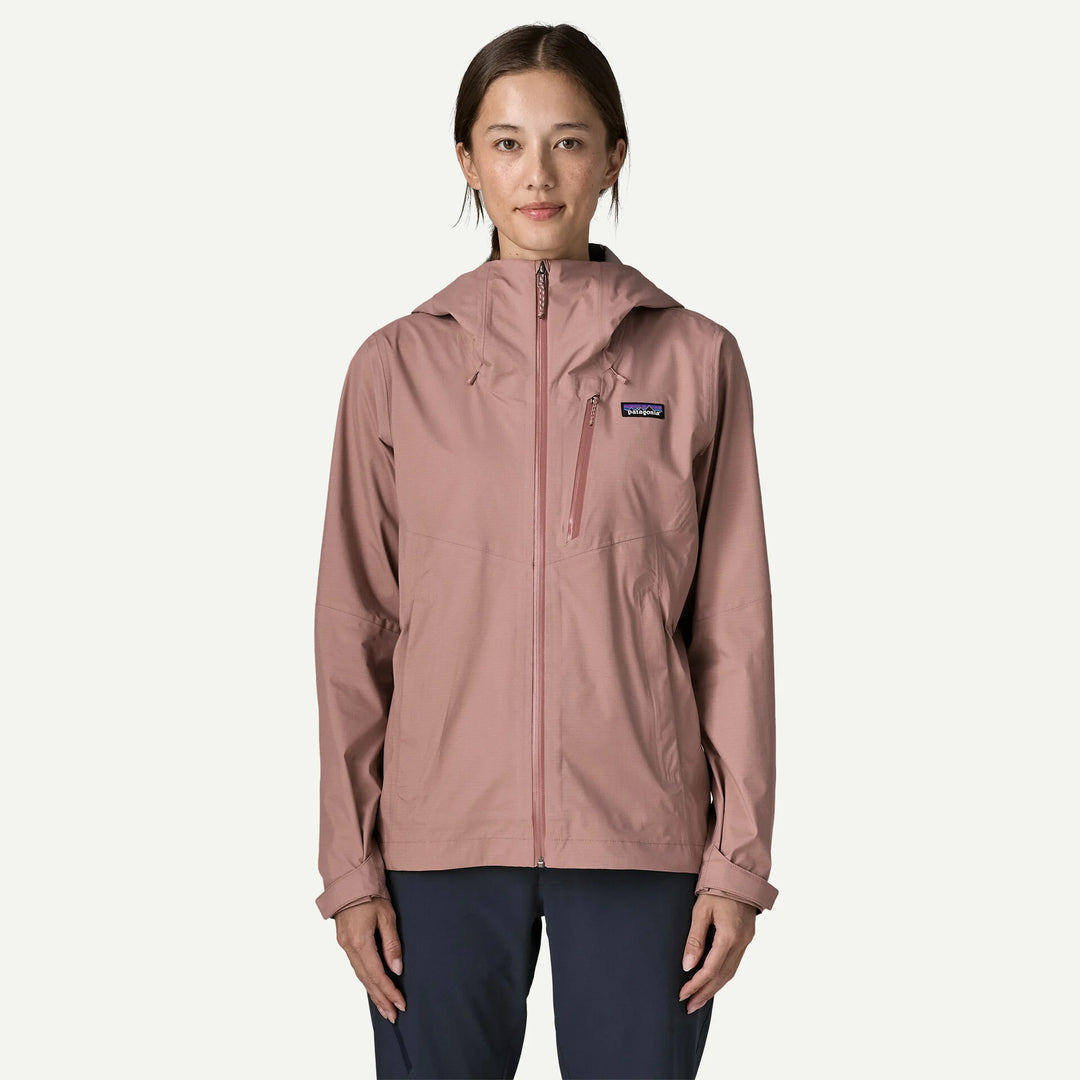 A woman wearing a Patagonia hooded jacket in dusty rose pink, paired with dark navy pants, standing against a light off-white background.