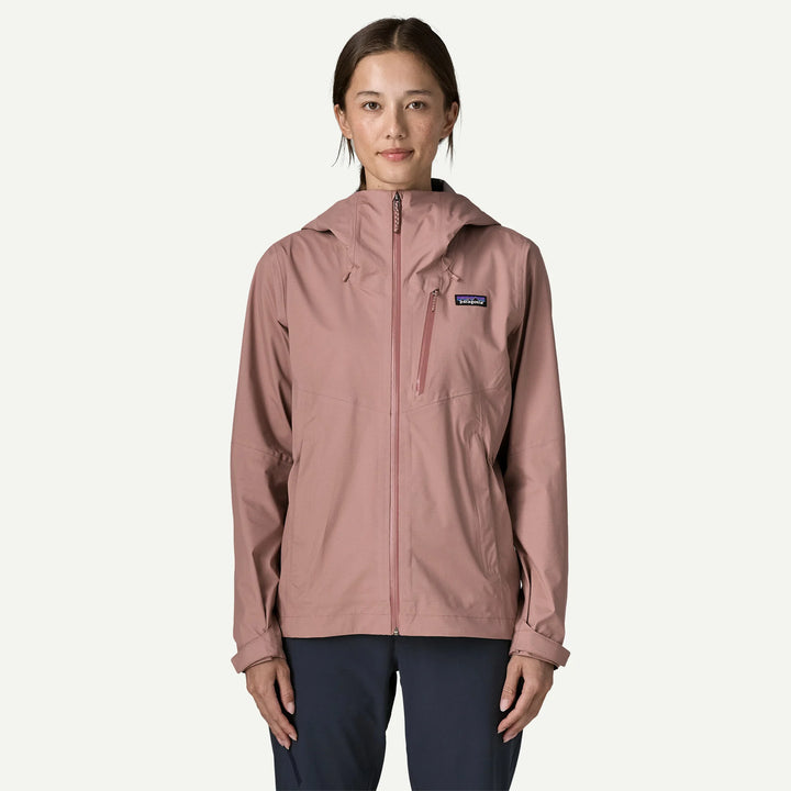 A woman wearing a Patagonia hooded jacket in dusty rose pink, paired with dark navy pants, standing against a light off-white background.