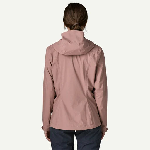 A woman viewed from the back wearing a dusty rose hooded jacket and dark navy pants against a clean off-white background.