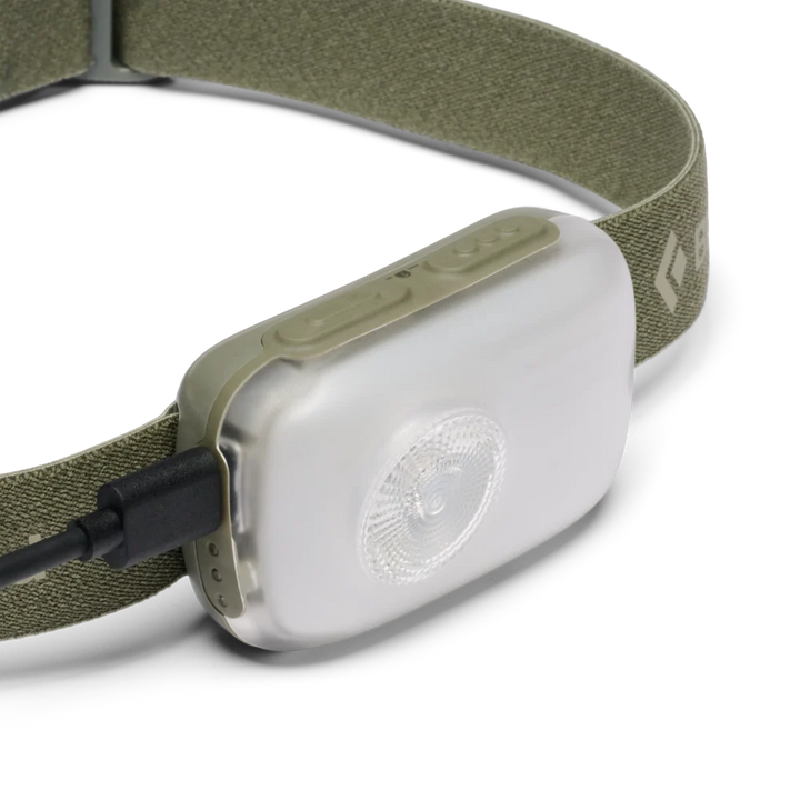A close-up of a headlamp with an olive green fabric strap and a white plastic body, shown on a clean white background with a USB charging cable plugged in.